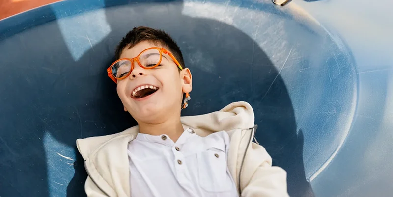 Ein lachender Junge mit orangefarbener Brille liegt entspannt auf einer blauen Rutsche und trägt ein weißes Shirt mit heller Jacke.