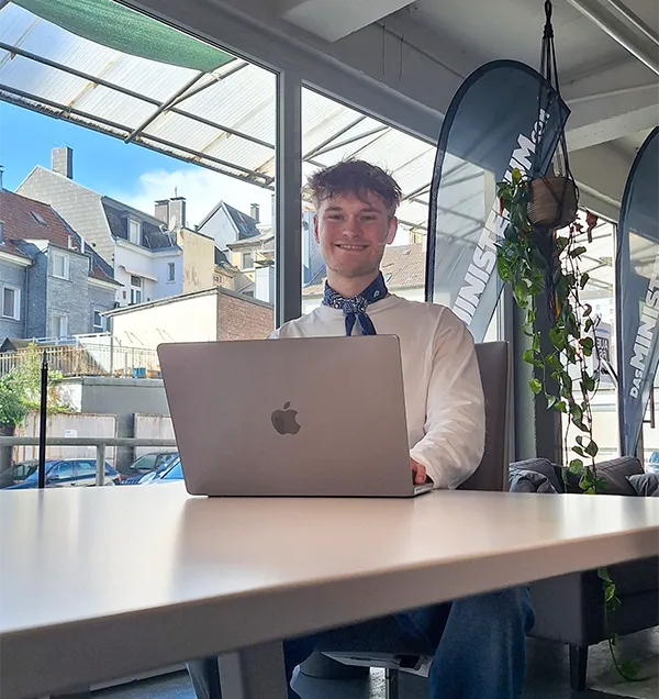 Junger Mann arbeitet mit Laptop an einem Tisch in einem hellen Büro mit großen Fenstern.